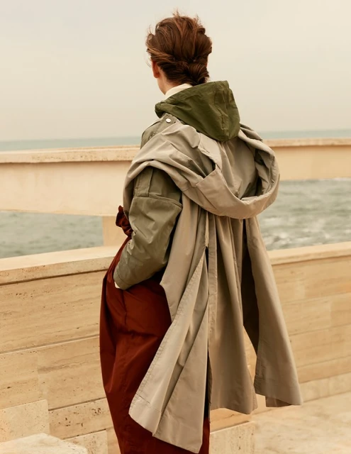 Woman in stylish outerwear overlooks the ocean on a cloudy day, standing by a light-colored wall, showcasing a blend of fashion and nature.