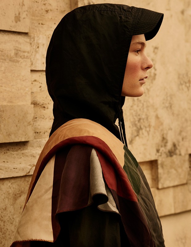Woman in profile wearing a black hooded cape with multicolored panel, set against a textured tan wall in a fashion editorial image.