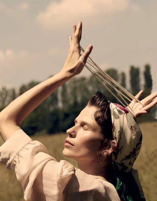 Woman in headscarf holds elastic bands over her head outdoors on sunny day, looking upward with trees and sky in the background.
