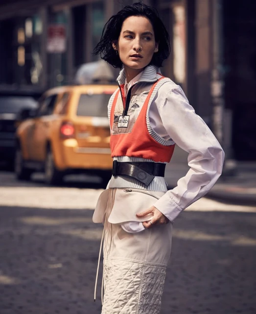 Stylish woman in orange vest and quilted skirt poses on city street with yellow taxi cab. Urban fashion editorial, daylight.