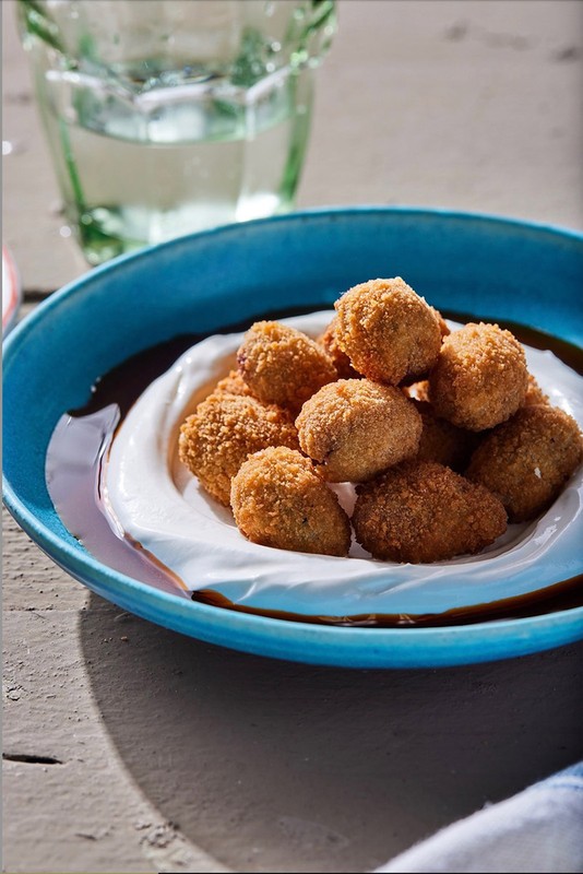 Elevated view of golden-brown arancini balls nestled on a white sauce bed inside a blue ceramic bowl, accompanied by a glass of water.