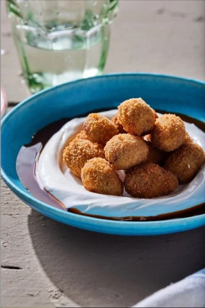Elevated view of golden-brown arancini balls nestled on a white sauce bed inside a blue ceramic bowl, accompanied by a glass of water.