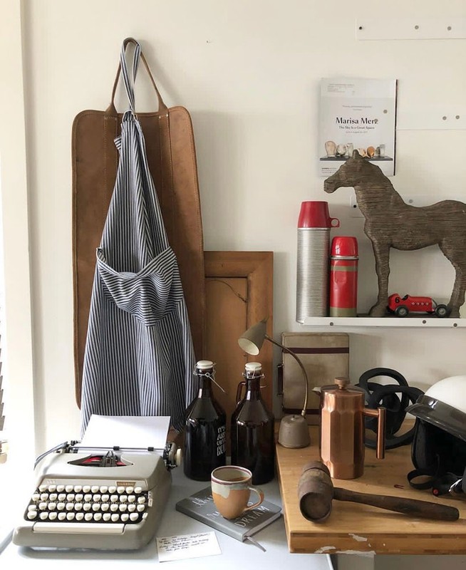 Still life composition with vintage typewriter, brown bottles, leather apron, and decorative objects against a neutral wall in a bright room.