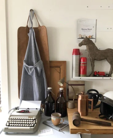 Still life composition with vintage typewriter, brown bottles, leather apron, and decorative objects against a neutral wall in a bright room.
