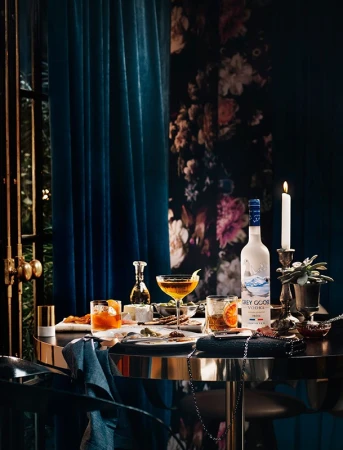 Grey Goose vodka bottle on a table with cocktails, snacks, and a floral wallpaper backdrop creating an inviting, sophisticated ambiance.