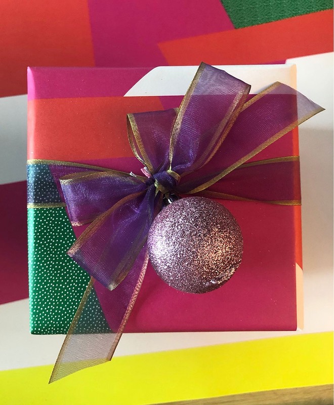 Festive gift box wrapped in colorful striped paper, tied with a purple sheer bow and decorative ornament, on a yellow background.