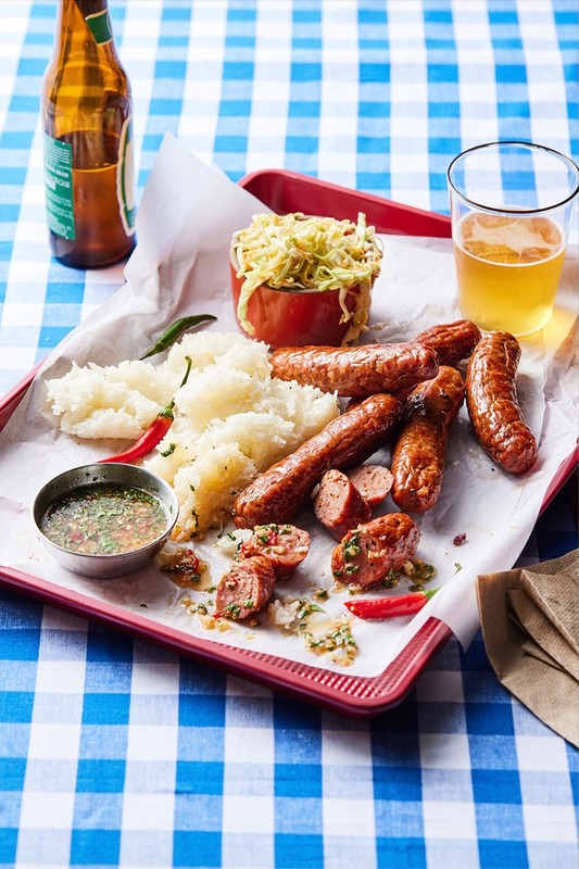 Overhead shot of sausages, rice, coleslaw, beer bottle, and beer glass on a serving tray with a blue checkered tablecloth.