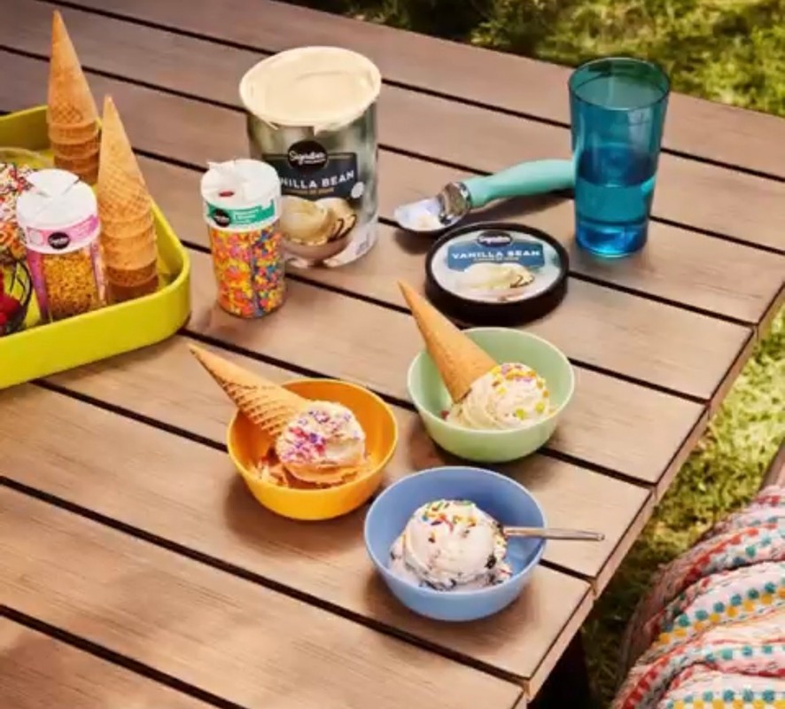 Ice cream cones in bowls and toppings arranged on a wooden table outdoors, creating a colorful dessert display.