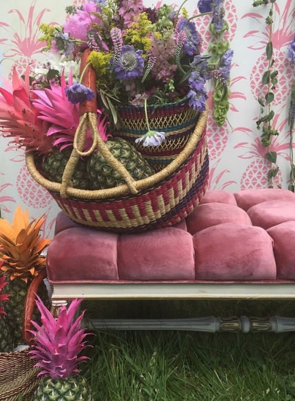 Close-up of a colorful floral arrangement and pineapples in a woven basket sitting on a pink tufted bench with a pineapple patterned wall.