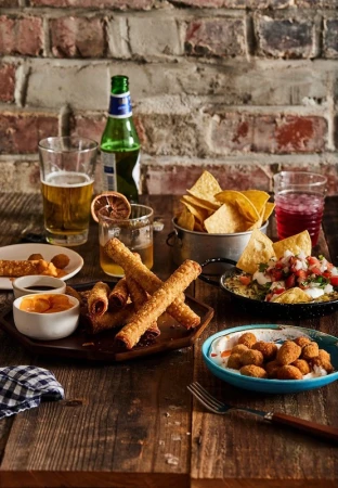 A table setting featuring a variety of appetizers, including fried snacks, chips, dips, and drinks, all arranged against a brick wall backdrop.