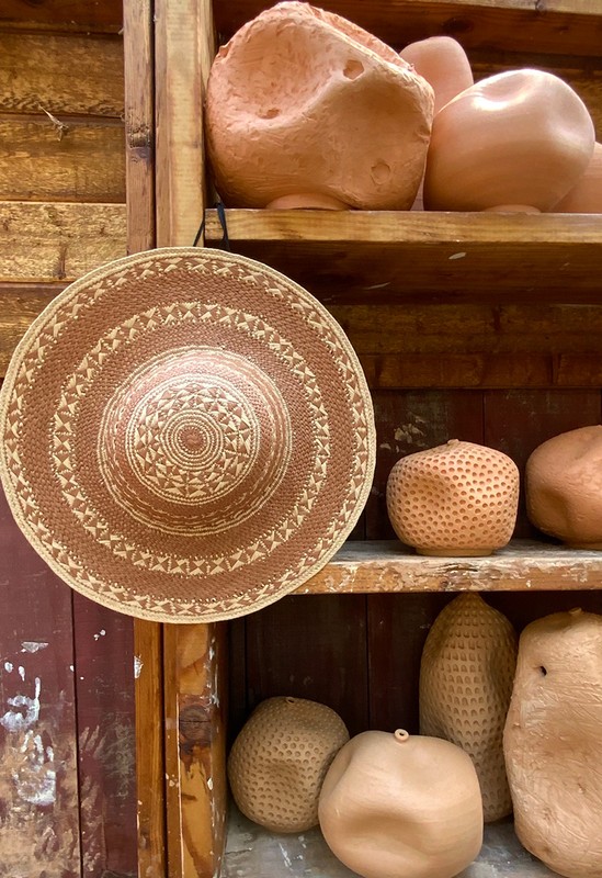 Close-up of a woven hat hanging on a wooden shelf next to a collection of bulbous pottery pieces in various sizes and shapes.