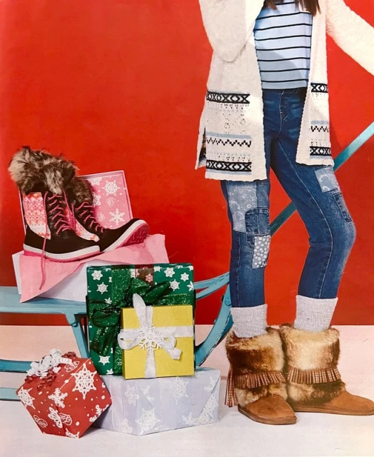 Young woman in casual winter clothing with patchwork jeans and fur boots posing amid a stack of wrapped Christmas gifts on a blue sled.
