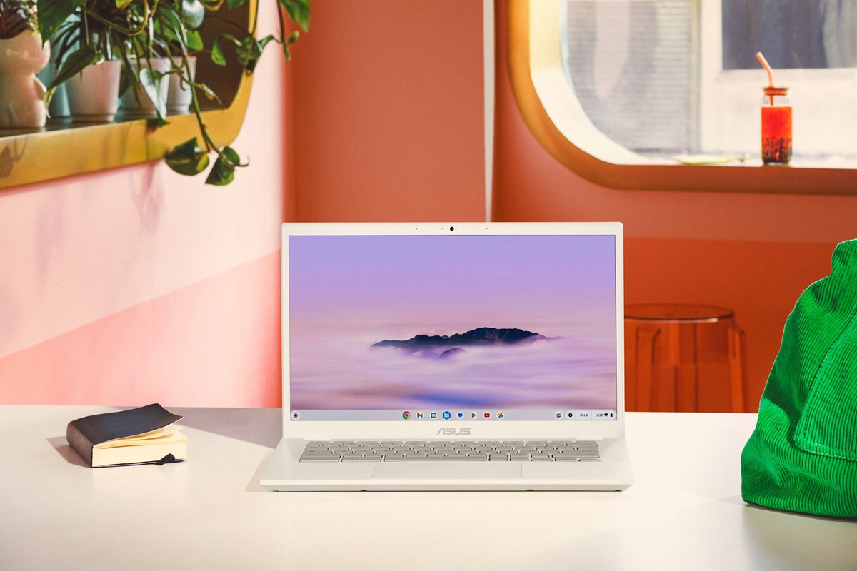 Modern white laptop on a white table with a pink wall background, a plant, a notebook, a drink, and a green bag.