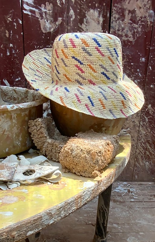 A colorful woven straw hat sits atop a wooden table with pottery tools, in an indoor pottery studio setting with natural lighting.