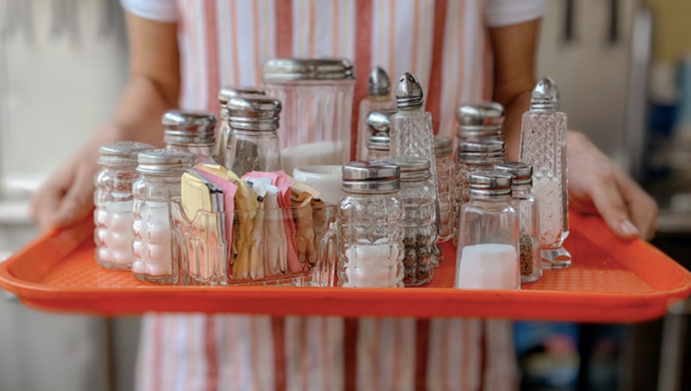 A person is carrying an orange tray filled with glass salt shakers and sugar packets with a striped background, showcasing a restaurant setting.