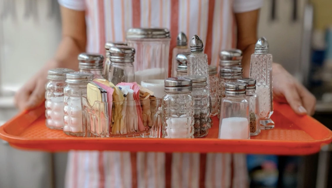 A person is carrying an orange tray filled with glass salt shakers and sugar packets with a striped background, showcasing a restaurant setting.