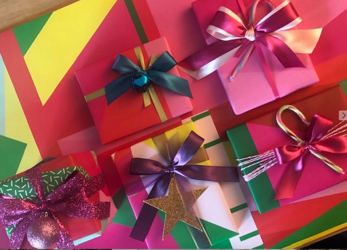 Collection of wrapped gifts in colorful paper with ribbons and bows of various styles on a vibrant, geometric background.
