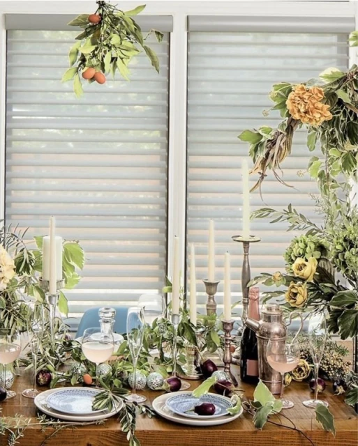 Elegant table setting with floral arrangements, candles, and drinks, featuring a wooden table and soft natural light from window blinds.