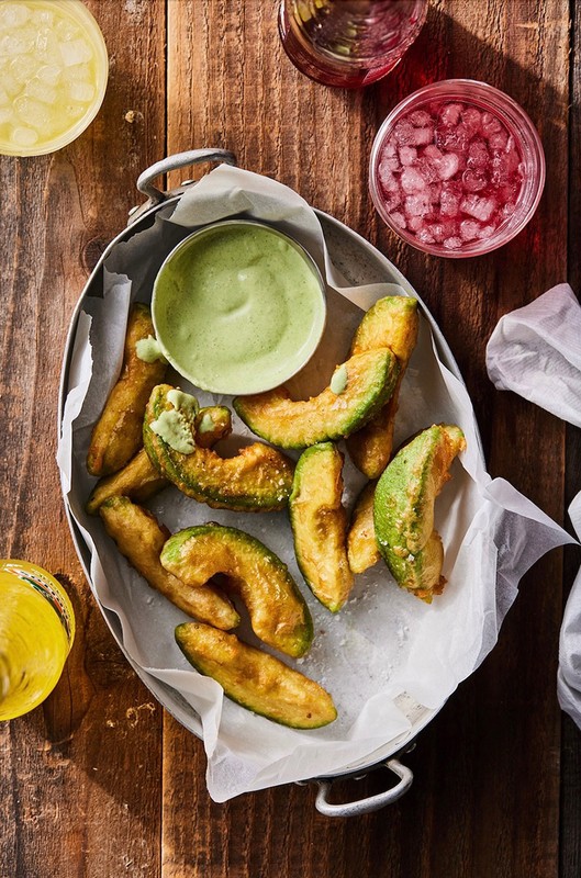 Overhead shot of fried avocado wedges and dipping sauce in a metal tray, set on a rustic wooden surface with drink garnishes.