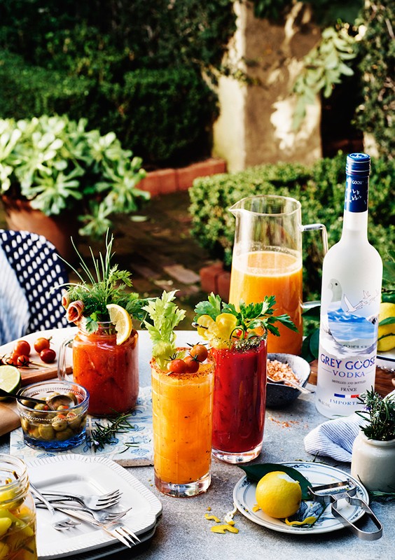 Outdoor brunch table with colorful cocktails, Grey Goose vodka, and assorted gourmet food items, plants, and garden background.