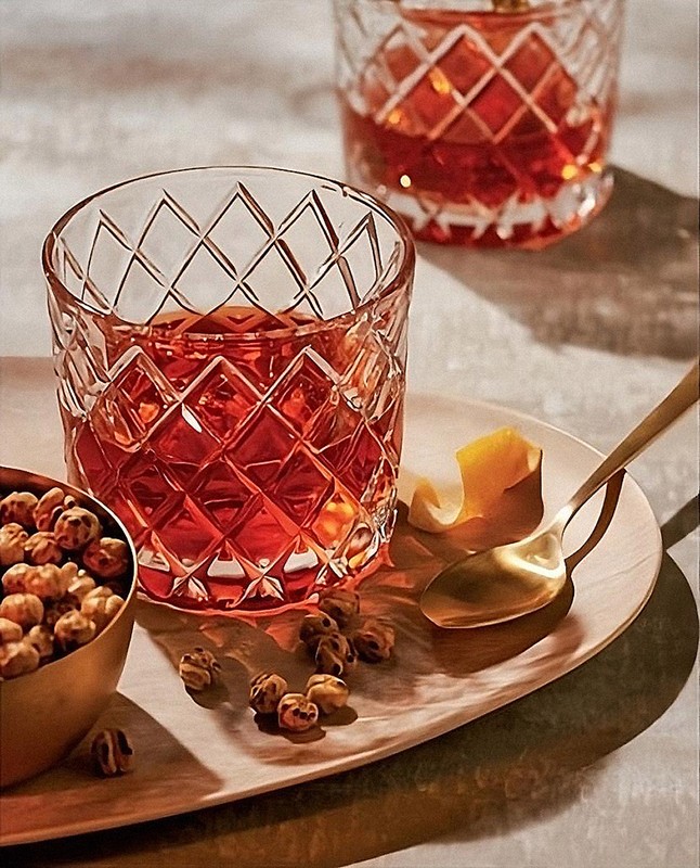 Two crystal glasses of amber cocktail with lemon peel on wood serving tray with bowl of chickpeas, set on neutral surface.