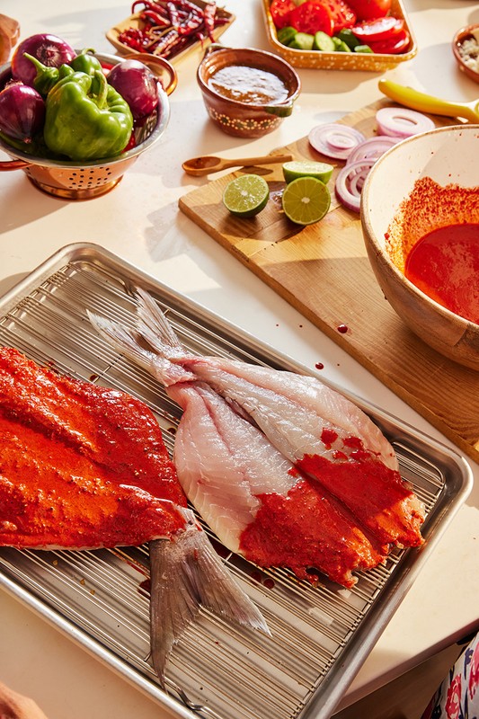Close-up of uncooked fish prepared with red sauce on a metal rack alongside tomatoes, onions, and peppers on a bright white surface.