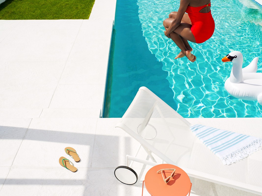 Woman in a red swimsuit jumps into a pool next to a swan floatie on a bright, sunny day with poolside lounge furniture and green grass.