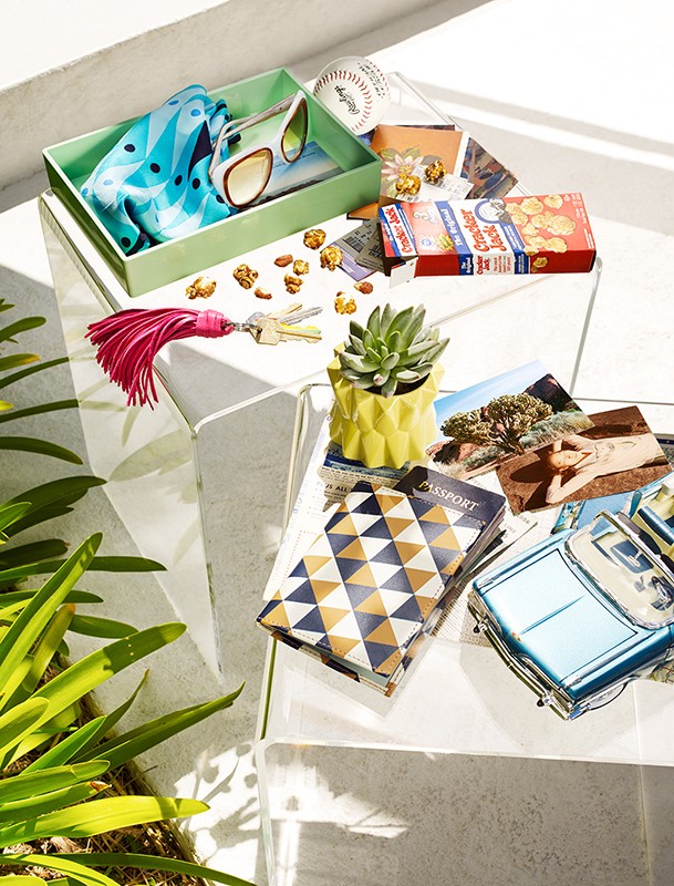 Still life of travel items, including a passport, postcards, sunglasses, and trinkets, arranged on a clear glass table outdoors.