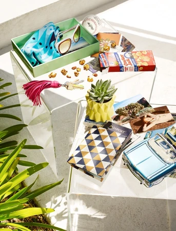 Still life of travel items, including a passport, postcards, sunglasses, and trinkets, arranged on a clear glass table outdoors.