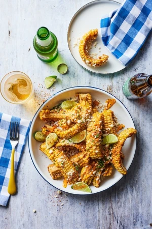 Overhead shot of elote on a white plate alongside beer bottles and lime wedges. A blue checkered napkin and yellow fork frame the food.