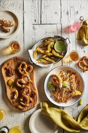 Overhead shot of pretzels, avocado fries, and drinks arranged on a rustic white wooden table, creating an inviting food spread.