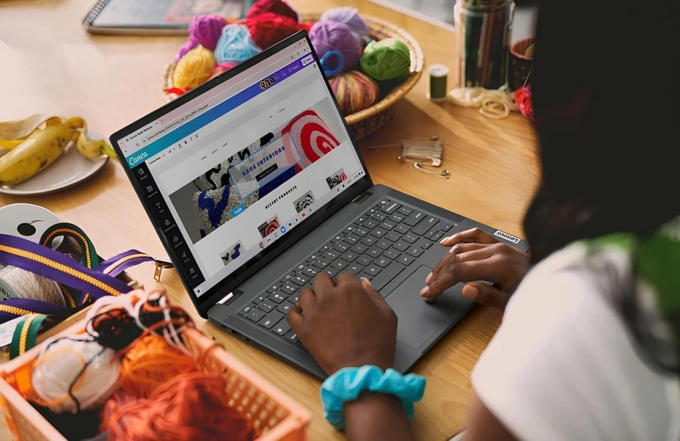 A person using a laptop on a wooden desk, surrounded by yarn and crafting supplies, including a partially peeled banana and ribbons.