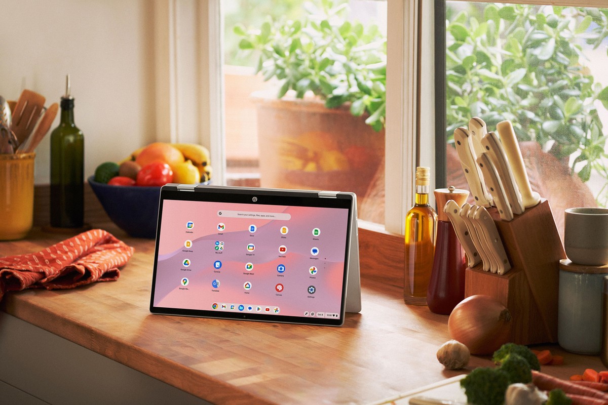 Chromebook tablet sits on a wooden kitchen counter next to a window, produce, knives, and cookware. Close-up shot with natural light.