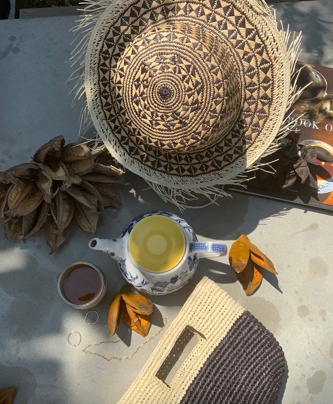 Overhead still life photo of tea service with a blue and white teapot, a cup of tea, a woven bag and hat on a concrete surface.