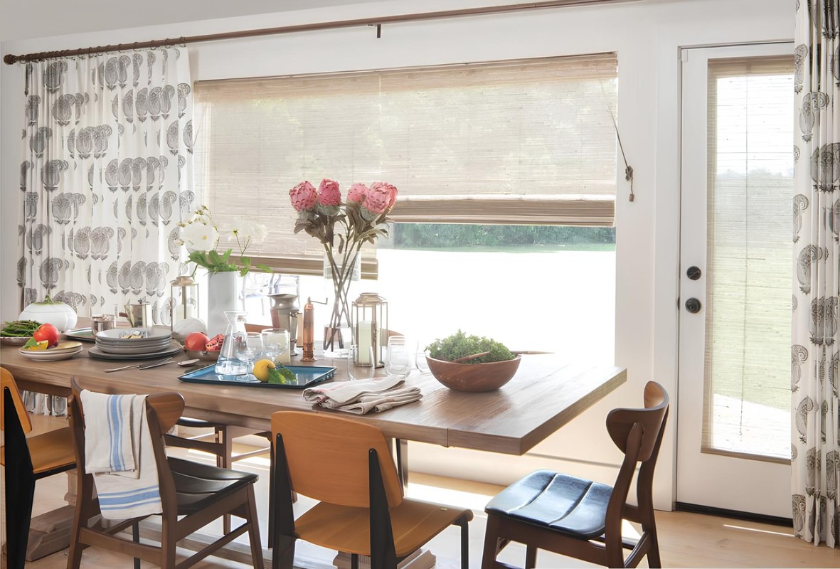 Dining room interior with a wooden table set for a meal, surrounded by chairs, flowers, and decorative curtains in natural light.