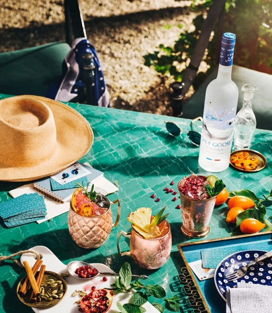 Outdoor table still life with Grey Goose vodka, copper mugs filled with mixed drinks, garnishes, fruit, sunglasses, and a hat.