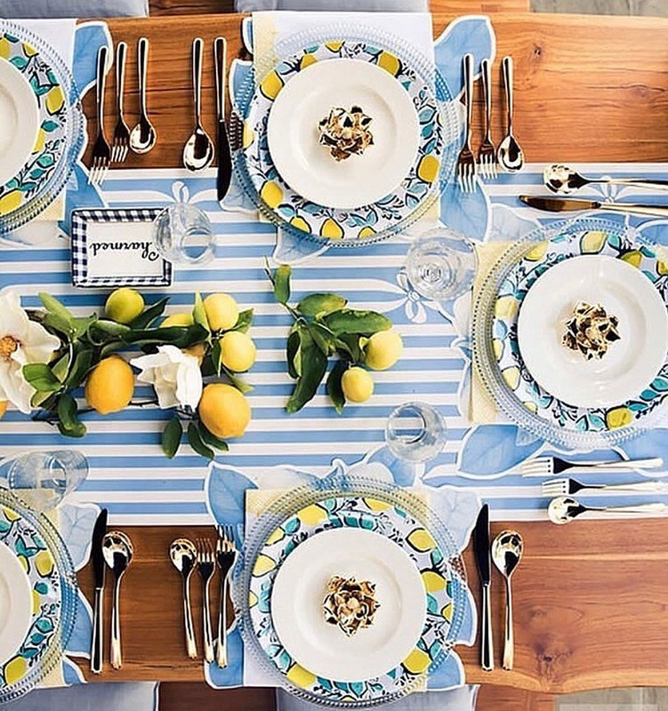 Overhead shot of a table setting with lemons, flowers, plates, silverware, and blue striped runner on wood surface for a stylish dining experience.
