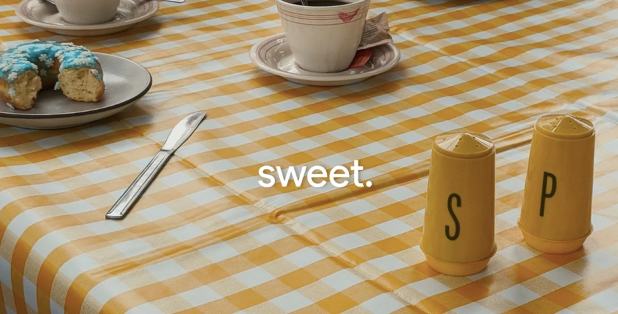 Overhead shot of a table with a yellow gingham tablecloth, featuring a donut, coffee cup, silverware, and salt and pepper shakers.
