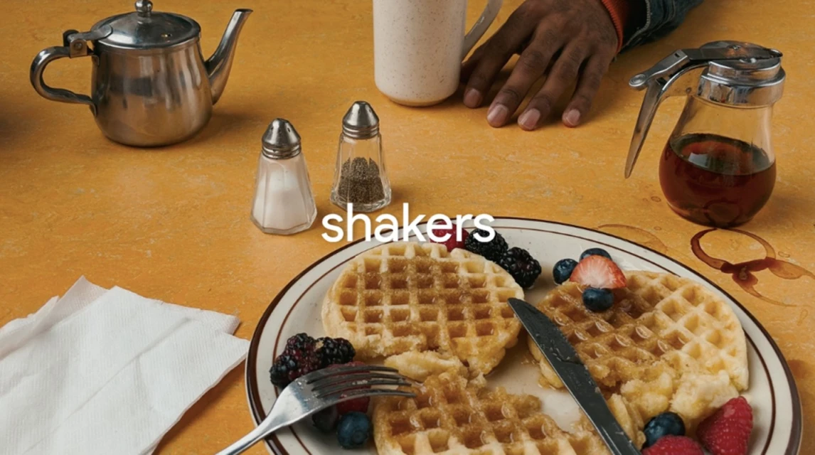Overhead shot of waffles with berries, syrup, salt & pepper shakers, silverware, and a beverage on a yellow table with a partial view of a hand.