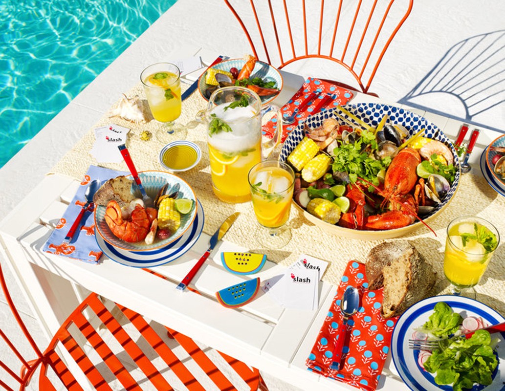 Outdoor seafood feast: lobster, shrimp, clams, corn, and drinks are arranged on a white table next to a pool with orange chairs.
