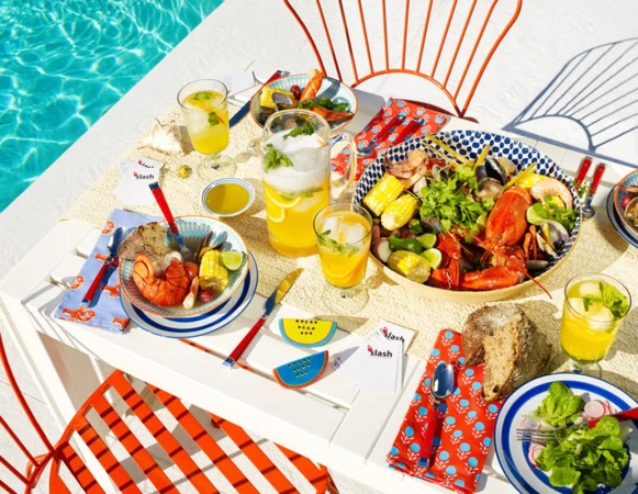 Outdoor seafood feast: lobster, shrimp, clams, corn, and drinks are arranged on a white table next to a pool with orange chairs.