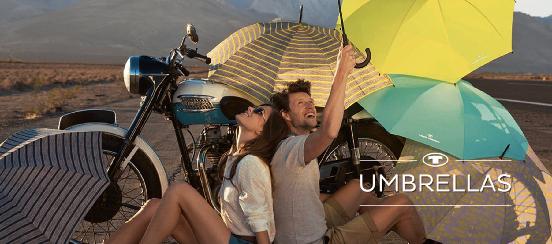 Couple sitting by motorcycle surrounded by umbrellas on an open road with mountain landscape in the background; product photography.