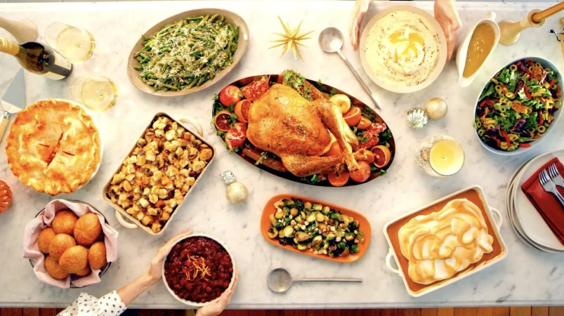 Overhead shot of a Thanksgiving or holiday feast on a white marble table with roast turkey, mashed potatoes, pie, salad, and side dishes.