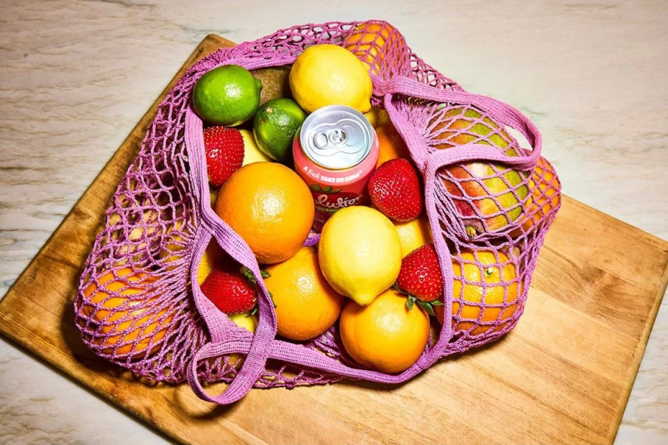 Overhead shot of a purple net bag overflowing with vibrant citrus fruits, strawberries, and a can of sparkling juice on a wood board.