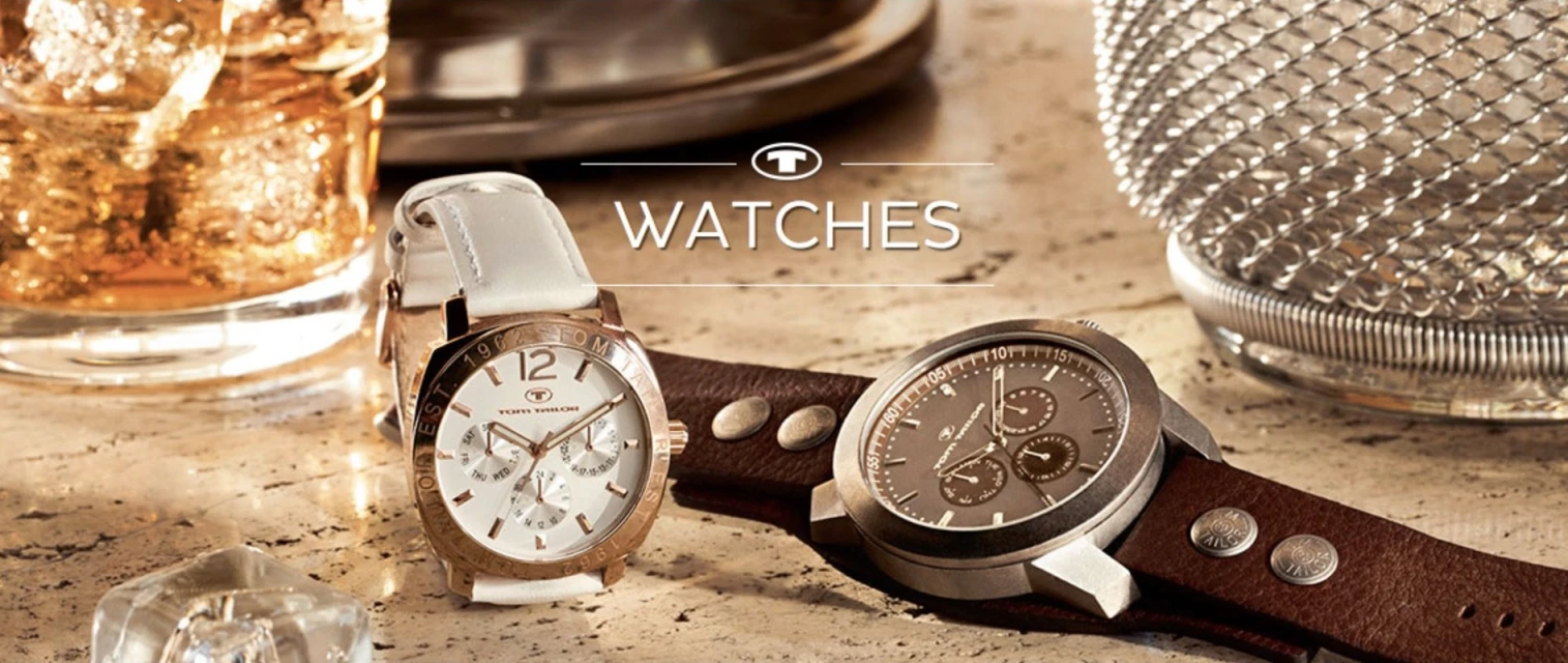 Two Tom Tailor wristwatches, one silver and one gold, are displayed on a marble surface next to an ice-filled glass and metal decor.