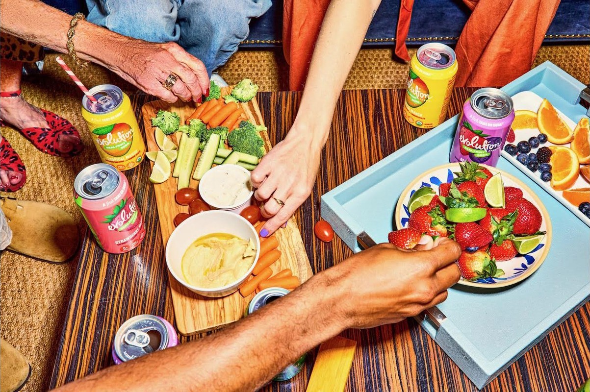 Overhead shot of several people enjoying appetizers and drinks, including carrots, celery, berries, and canned beverages.