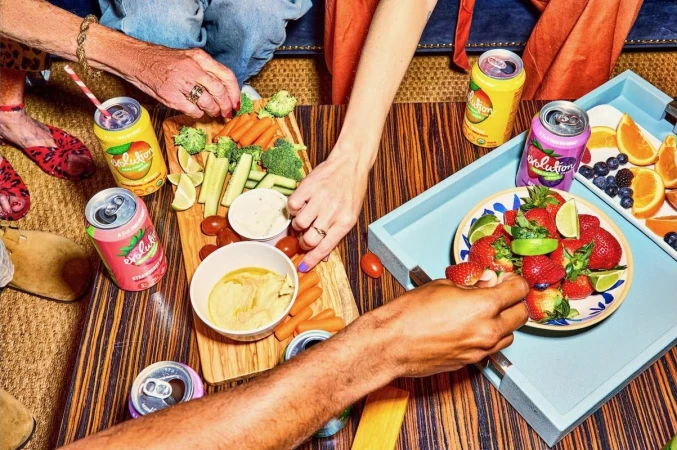 Overhead shot of several people enjoying appetizers and drinks, including carrots, celery, berries, and canned beverages.