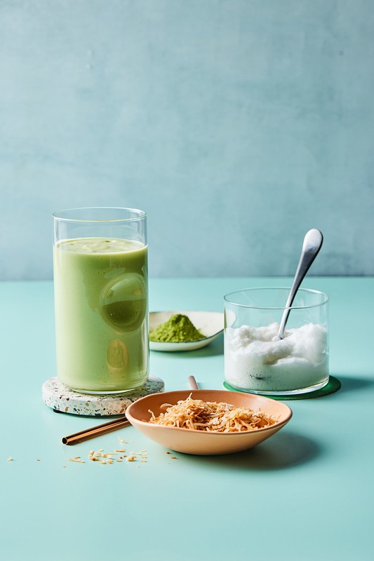 A glass of green matcha smoothie with a bowl of toasted coconut flakes, matcha powder, and shaved ice on a blue table.
