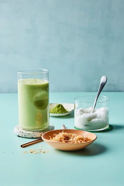A glass of green matcha smoothie with a bowl of toasted coconut flakes, matcha powder, and shaved ice on a blue table.