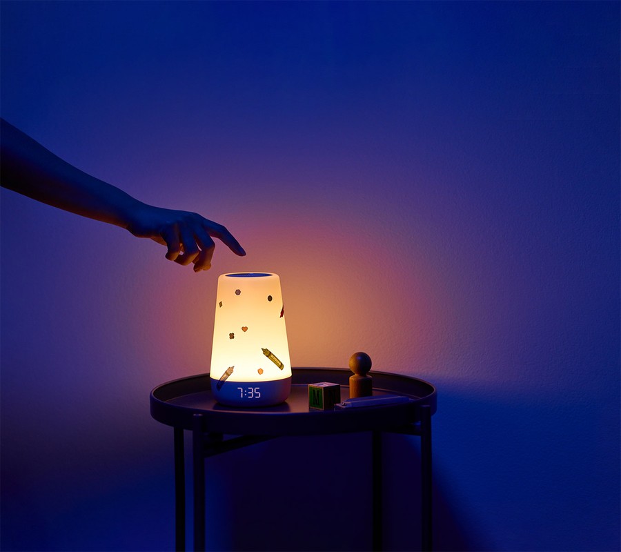 Illuminated white bedside lamp with colorful crayon pattern on a round black table with a hand reaching for the top.